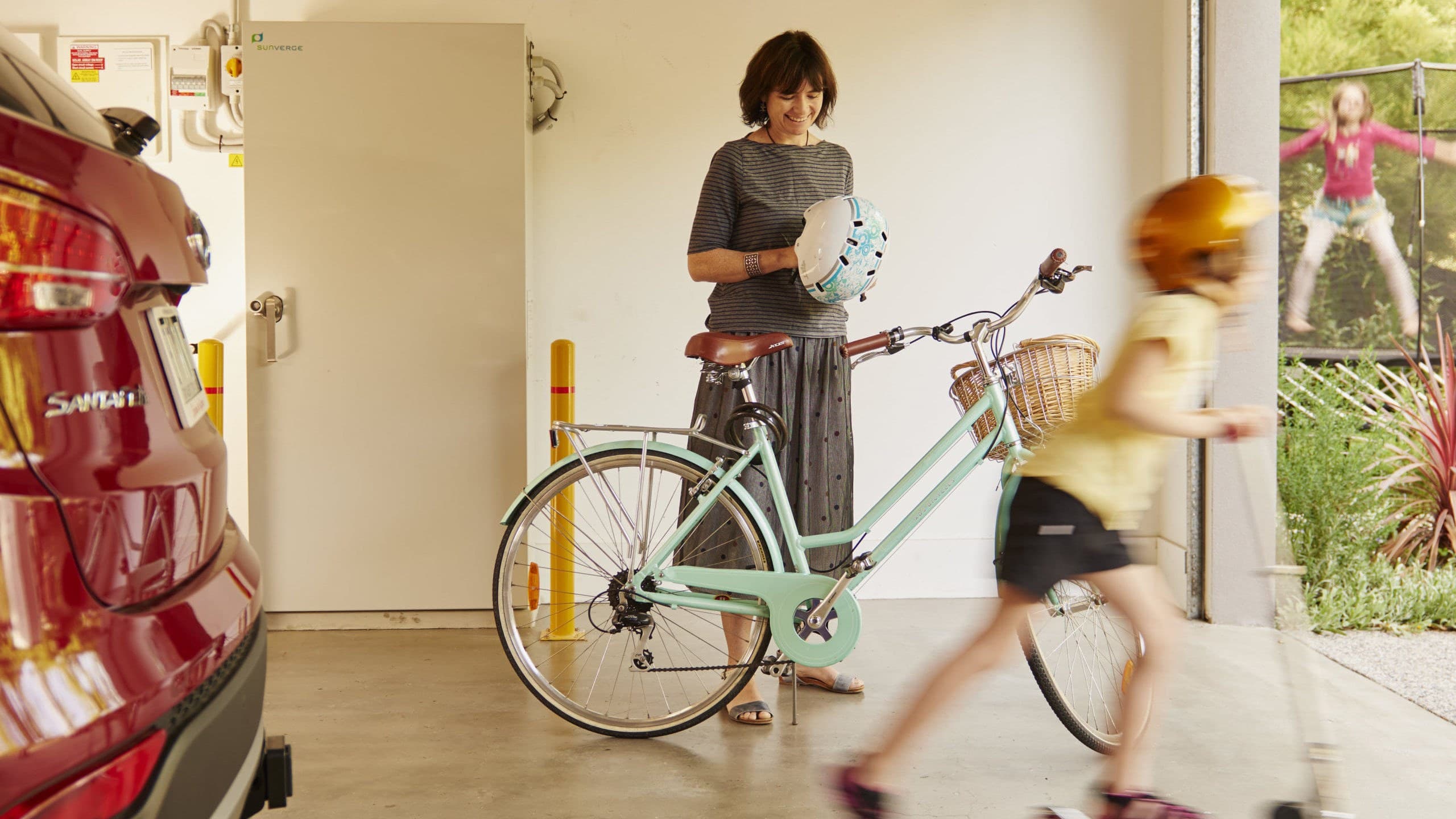 Woman at her home putting on helmet in front of bike in garage with young boy riding out on scooter